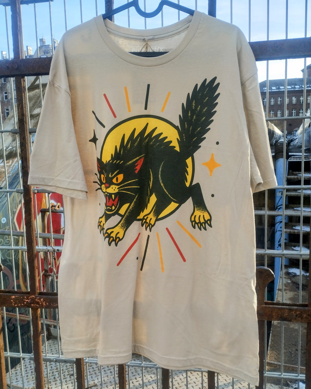 Beige t-shirt with a graphic of a black panther on a yellow circle, hanging against a metal fence.