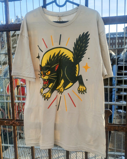 Beige t-shirt with a graphic of a black panther on a yellow circle, hanging against a metal fence.
