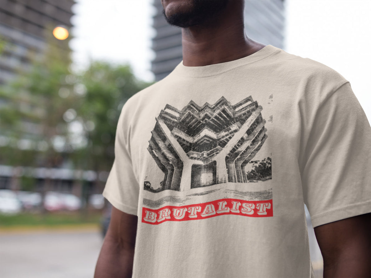 A man wearing a beige graphic t-shirt with a black geometric art print and a red banner reading 'BRUTALIST'. The background is a blurred outdoor city street scene.