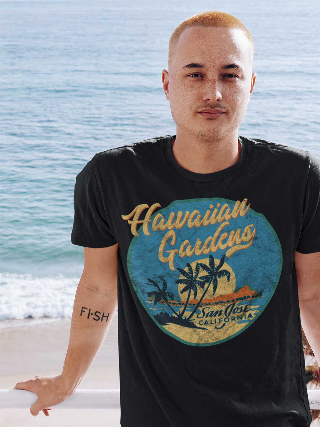 Man wearing a black t-shirt with 'Hawaiian Gardens' design by the ocean