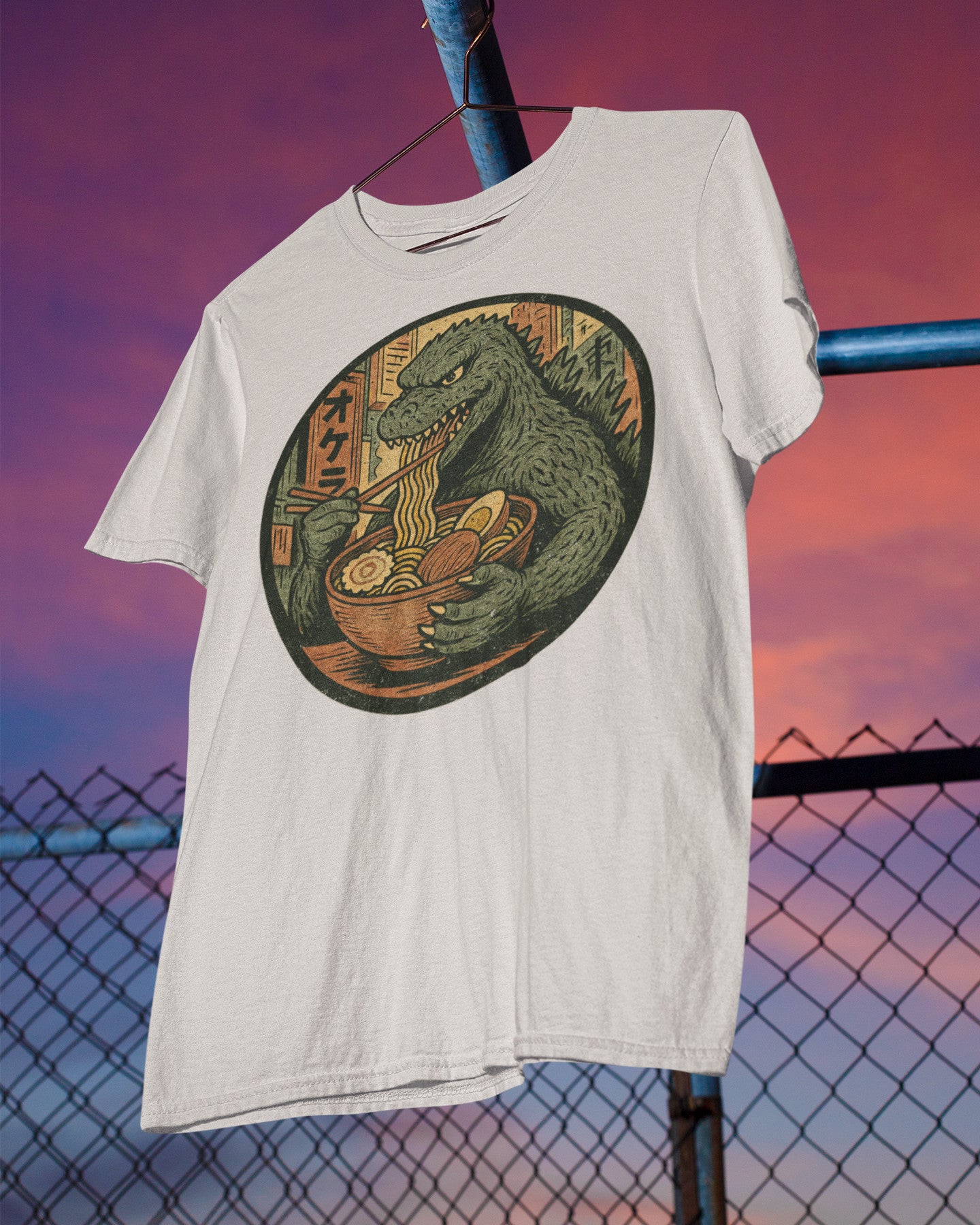 White t-shirt with Godzilla eating ramen graphic against a colorful sky.