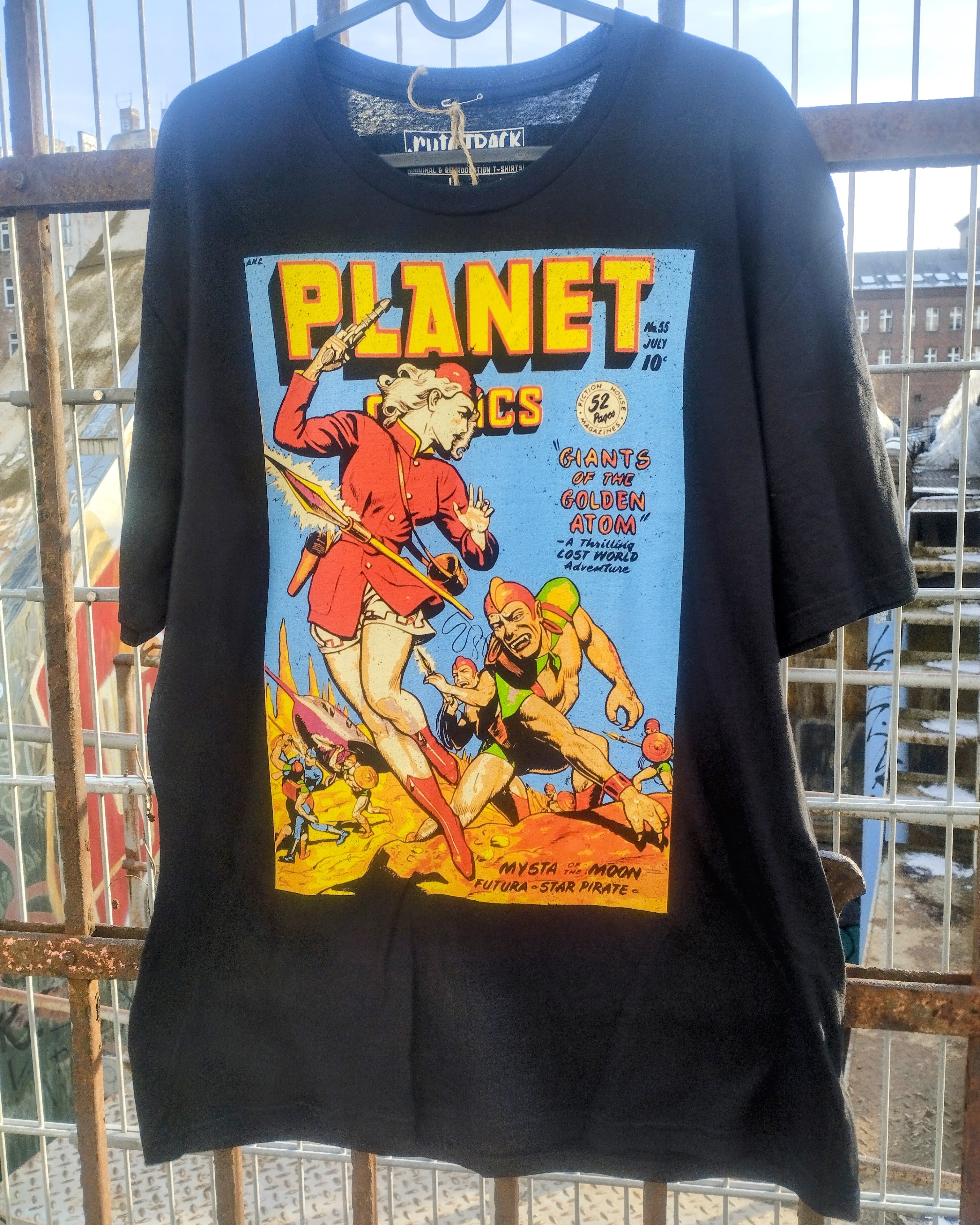 Black t-shirt with a graphic design of a comic book cover featuring a warrior and a creature, hanging on a metal rack.