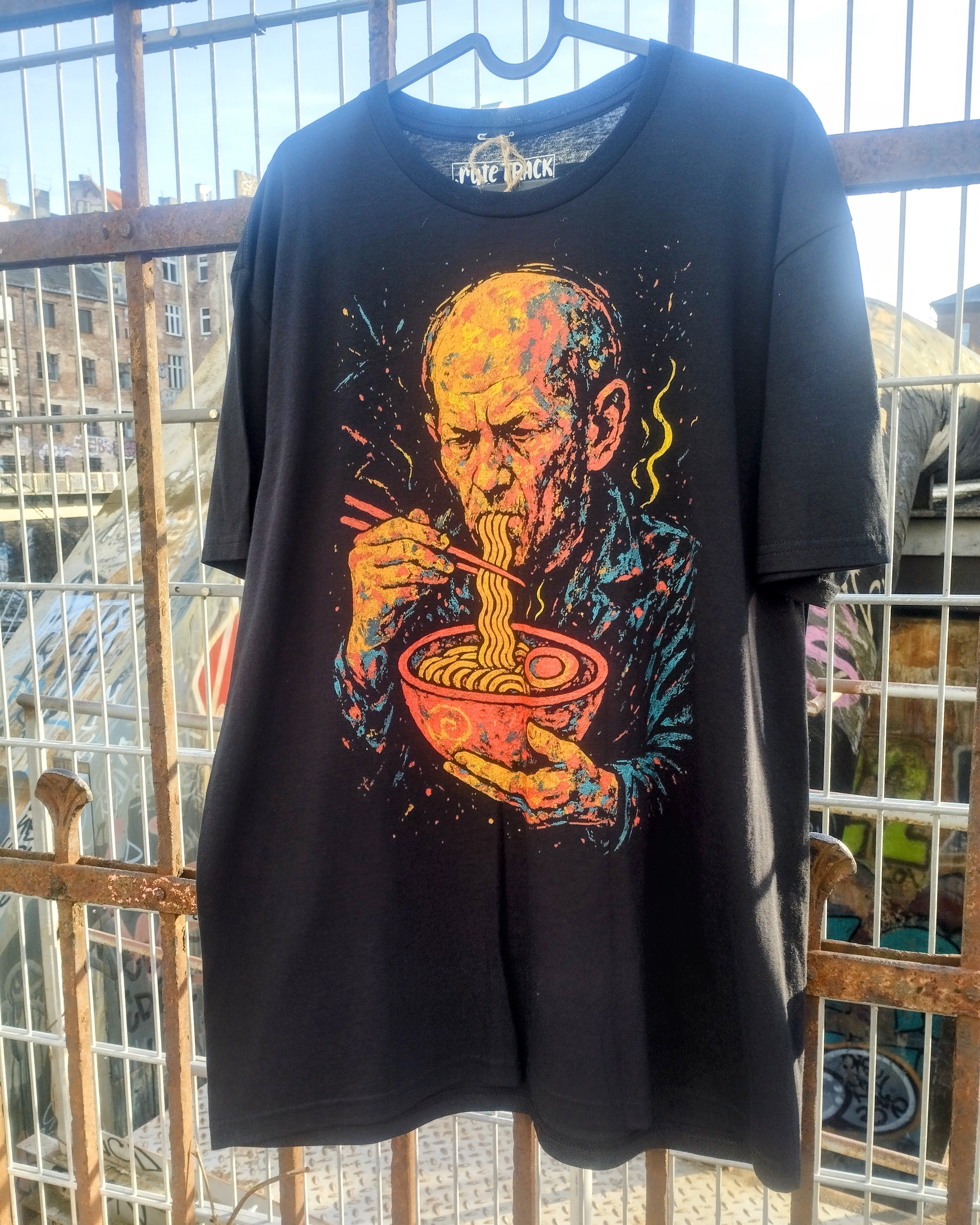 Black t-shirt with a colorful graphic of a man eating noodles, hanging against a metal fence.