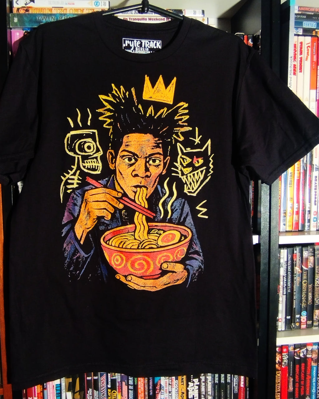 Black t-shirt with a graphic design of a person eating ramen noodles against a book case.