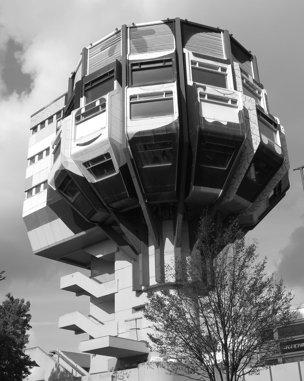 Black and white architectural photograph of a highly sculptural, multi-level modern building with cantilevered sections and a staircase base, set against a cloudy sky with trees in the foreground. By Roger Cowell 2026