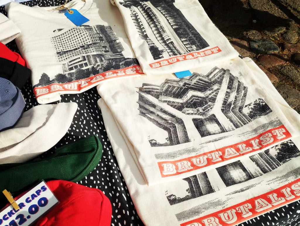 A stack of off-white graphic T-shirts laid out on a black-and-white patterned cloth, featuring black architectural Brutalist prints and red text reading 'BRUTALIST'. A small price tag and other folded garments are partially visible around the edges.