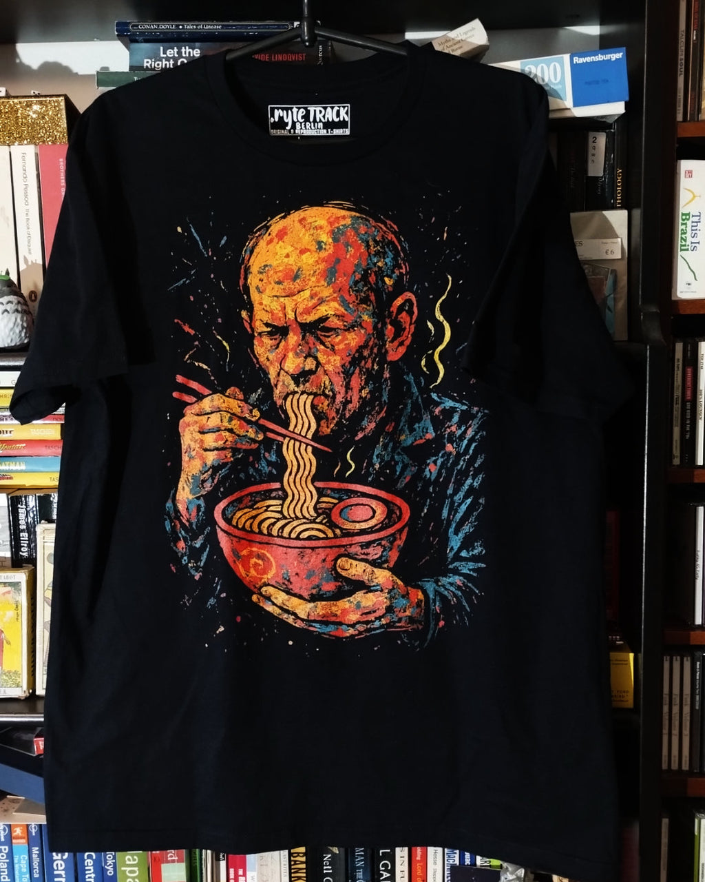 Black t-shirt with a graphic of a man eating noodles, displayed against a bookshelf background.