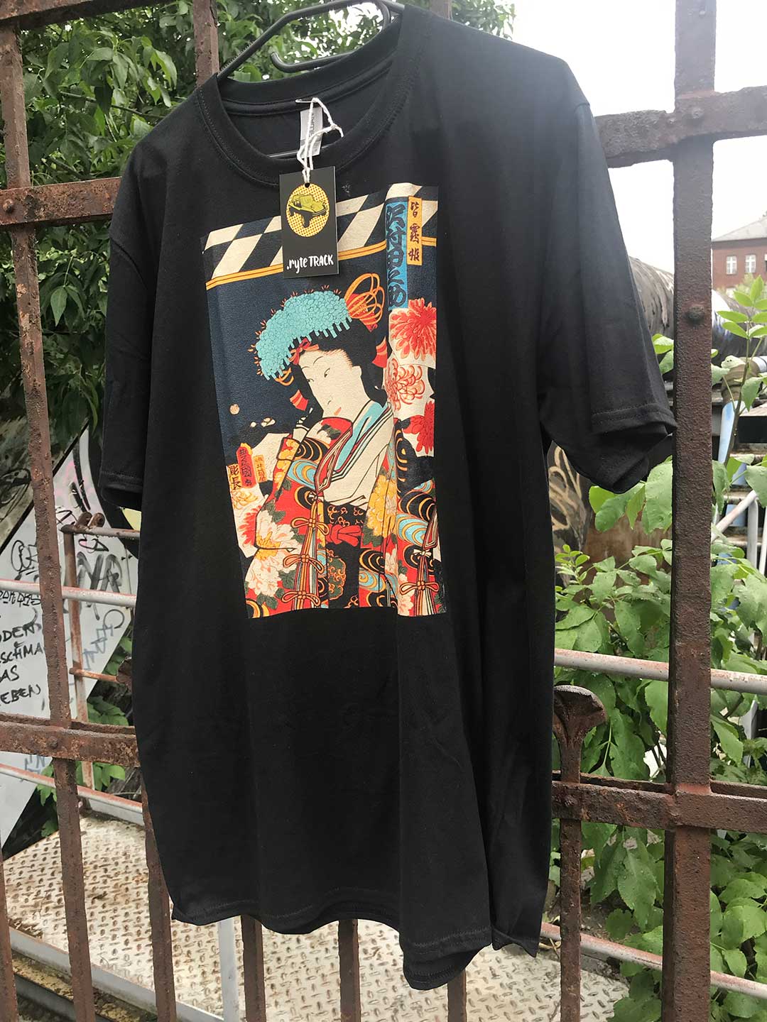 Black t-shirt with colorful Japanese graphic design hanging on a rusted metal fence.