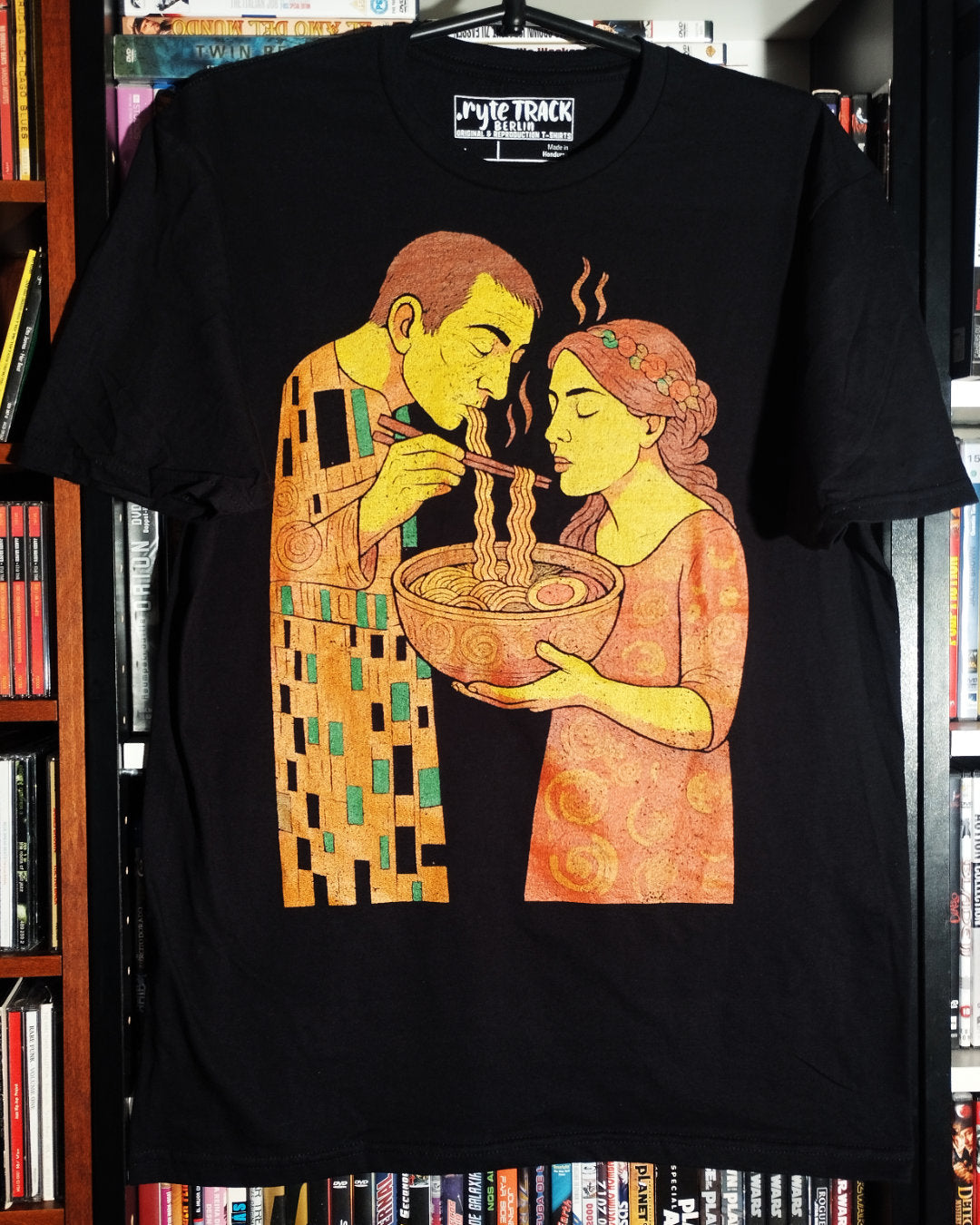 Black t-shirt with a stylized illustration of two people sharing a bowl of soup, hanging against a bookshelf background.