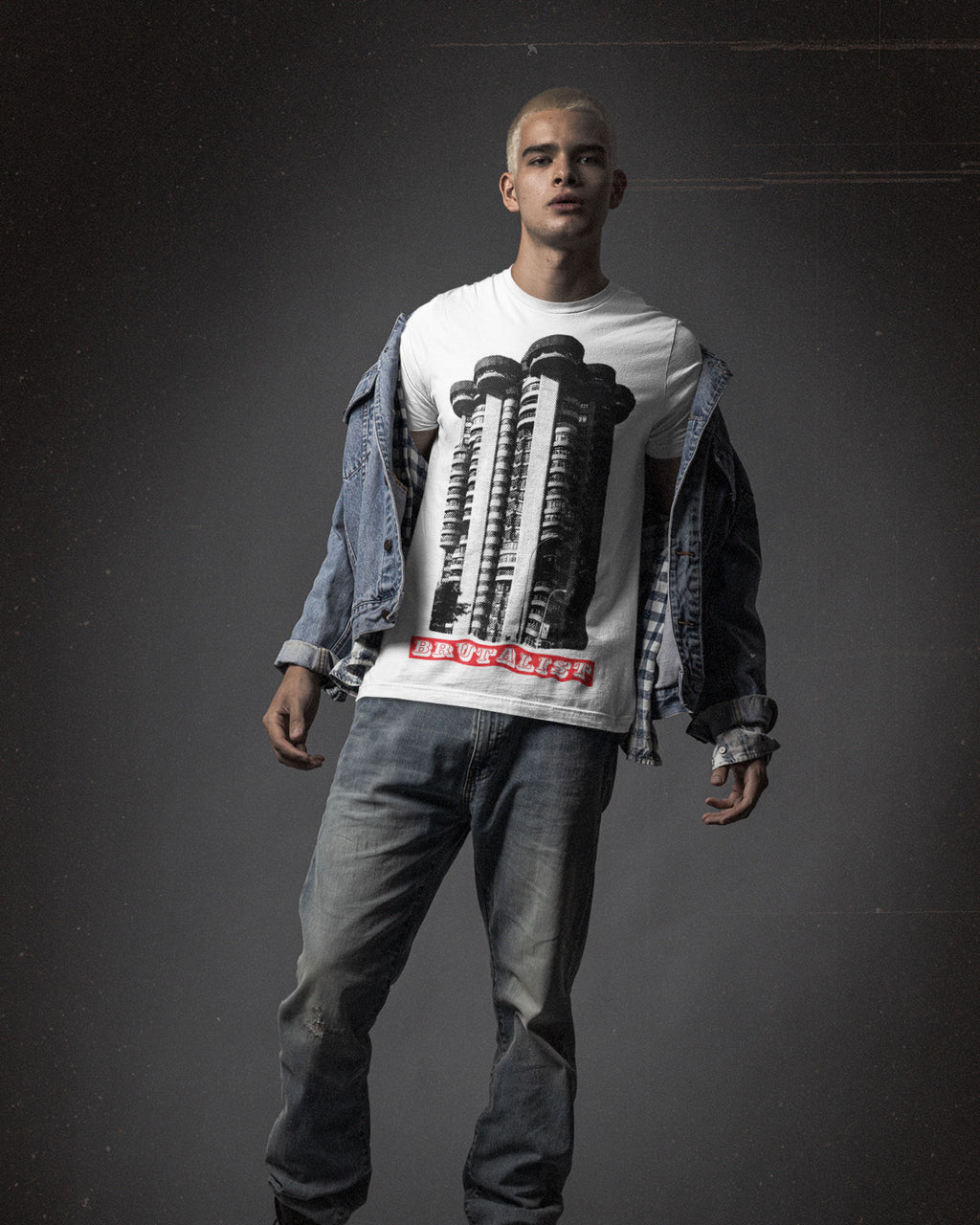 A man modeling a white graphic t-shirt with a black and red brutalist design, styled with an open blue denim jacket and gray jeans against a dark studio background.