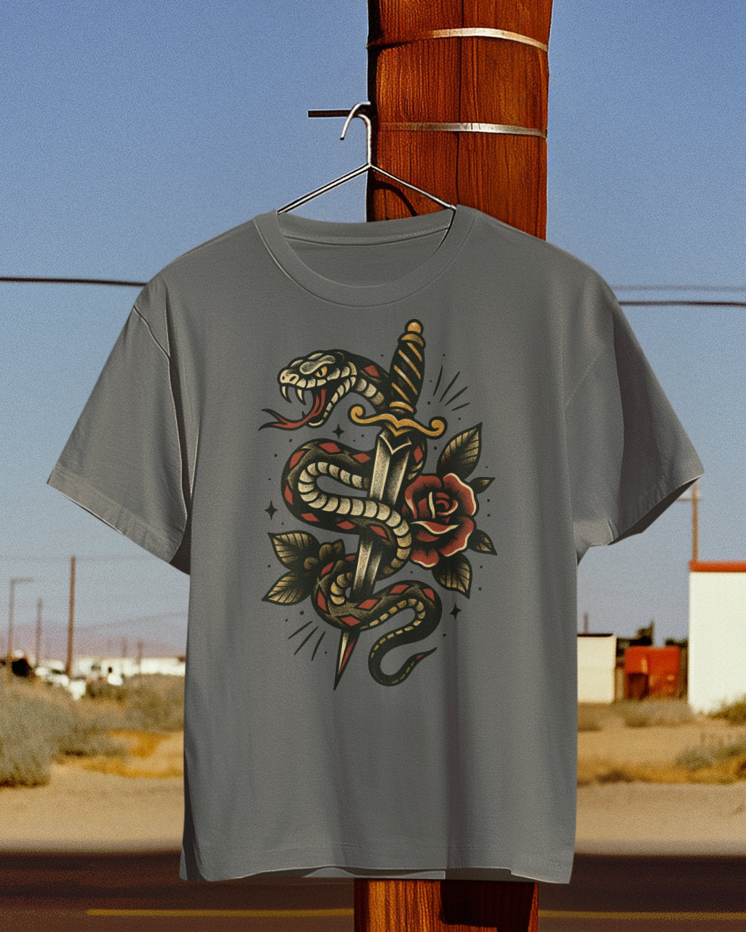 Gray t-shirt with snake and rose design hanging on a wooden post against a desert landscape.