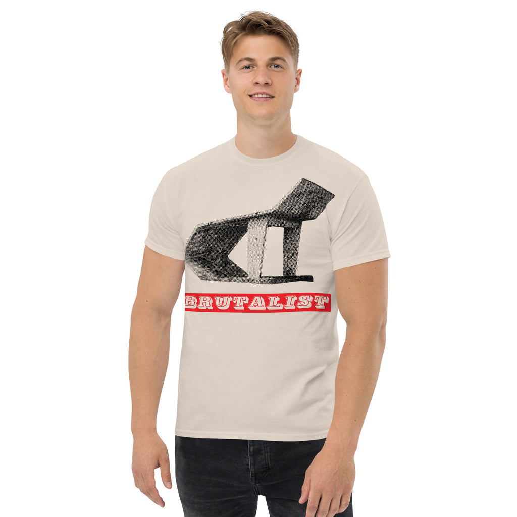 A man wearing a light beige crewneck t-shirt with a graphic of a Soviet era bus stop and the word 'BRUTALIST' in red, paired with dark jeans, against a plain white background.