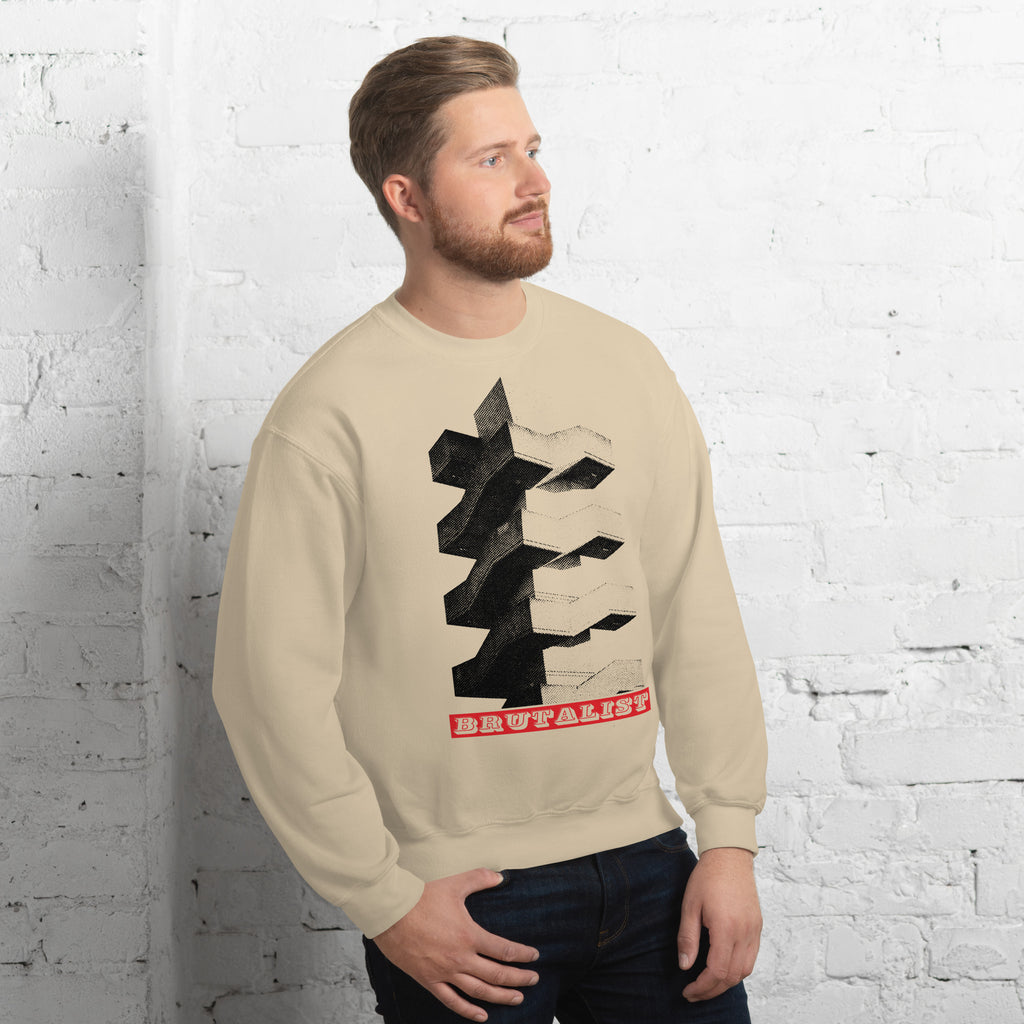 A man wearing a beige crewneck sweatshirt with a black and red graphic print, standing against a white brick wall.