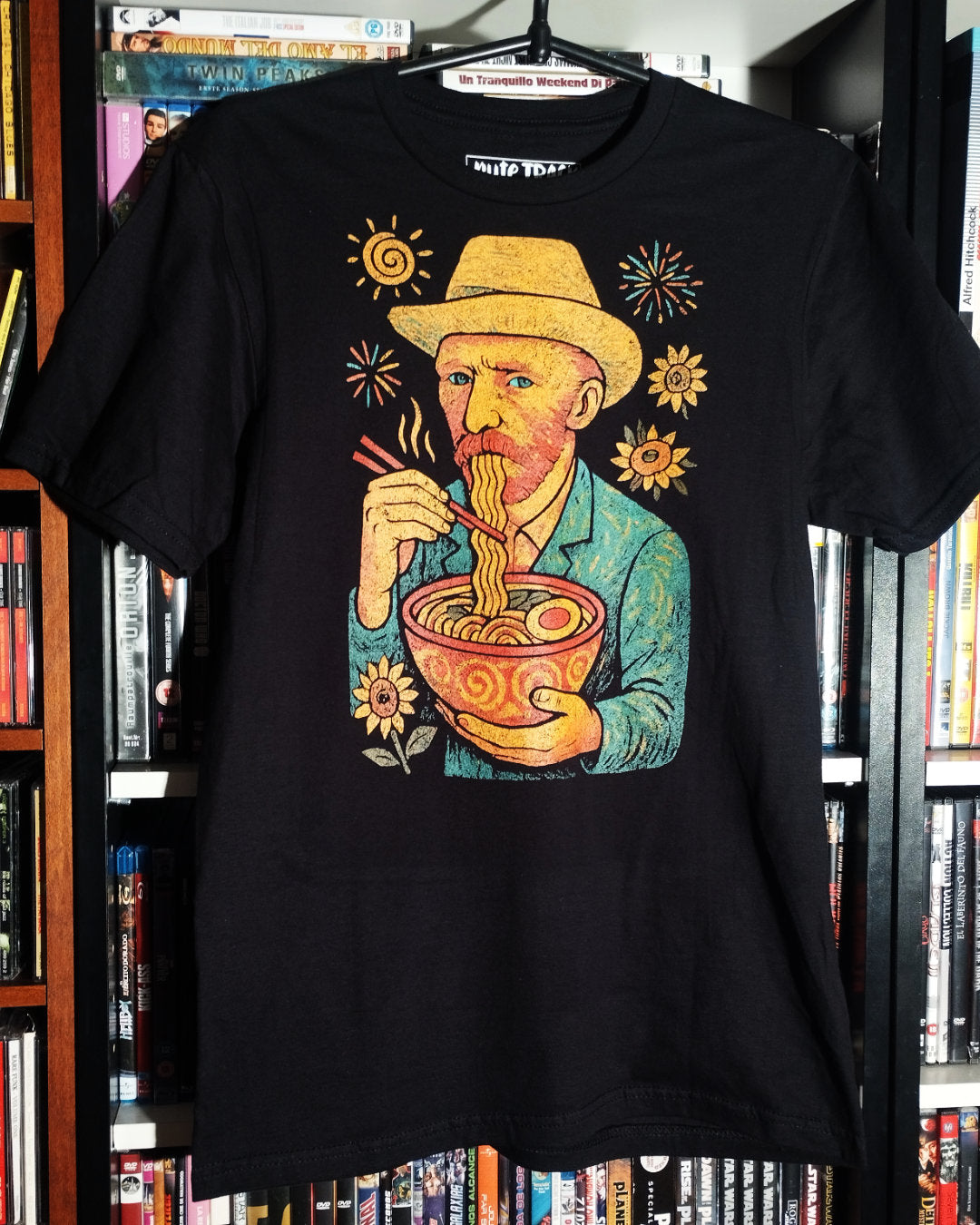 Black t-shirt with a colorful graphic of Vincent Van Gogh eating ramen, hanging in front of a bookshelf.